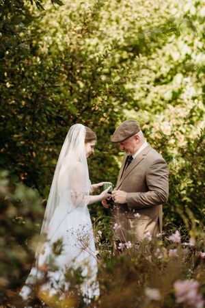 Bride in white gown and veil shares a tender moment with an older gentleman in a lush garden setting.