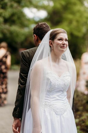 Bride in lace gown and veil smiles softly while holding grooms hand outdoors before ceremony.