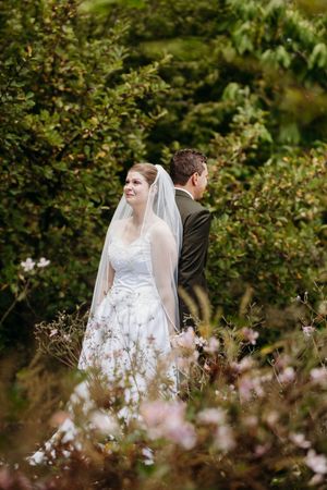 Bride in white wedding gown and veil stands beside groom among blooming flowers in a green garden.