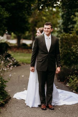Smiling groom in dark suit stands on garden path as bride in white gown peeks behind him.