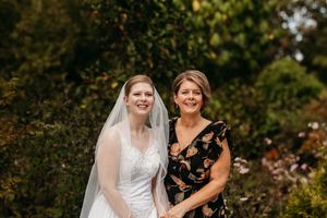 Radiant bride in white lace gown and veil poses with smiling woman in floral dress outdoors.