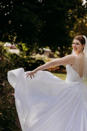 Bride in elegant white gown twirls her dress in a sunlit garden, smiling over her shoulder.