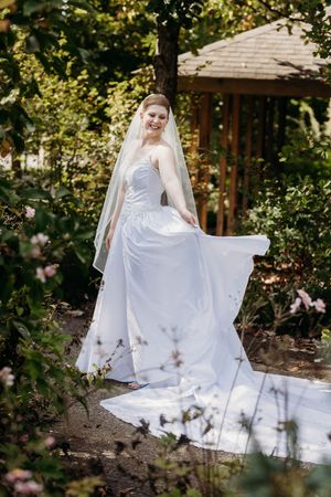 Bride in flowing white wedding gown and veil lifts her skirt while standing in a garden path.