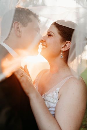 Bride and groom share a romantic moment under a veil with golden sunset light glowing between them.