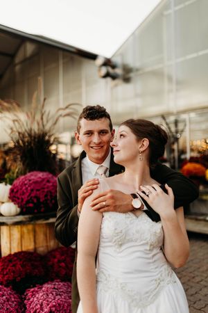 Groom embraces bride from behind as they pose outdoors surrounded by vibrant purple and red mums.