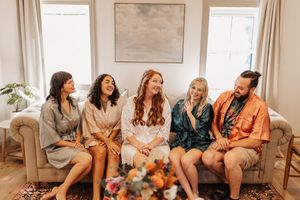 Bride and wedding party laugh together on couch in silk robes.