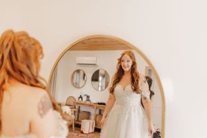 Redheaded bride admires herself in large round mirror in bridal suite.