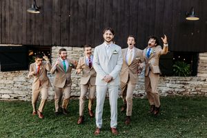 Groom in light gray suit stands with groomsmen in tan suits celebrating outdoors near stone wall and barn.