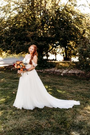 Bride in off-shoulder white tulle gown holding vibrant orange and red bouquet in sunlit garden setting.
