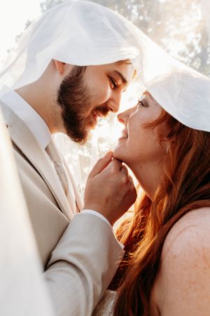 Bride and groom sharing intimate moment under wedding veil in warm golden sunlight close-up portrait.