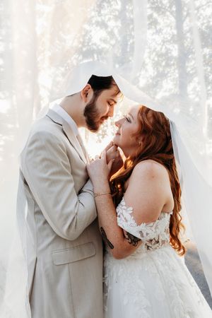 Bride and groom holding hands under flowing white veil in sun-drenched outdoor wedding portrait.