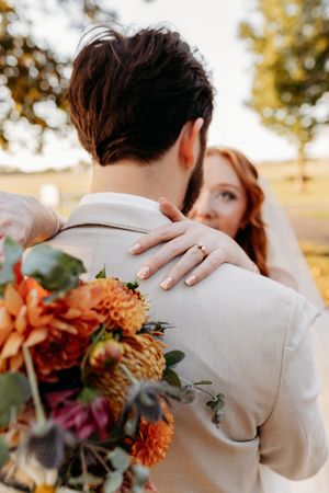 Bride embracing groom from behind holding orange dahlia bouquet with wedding ring visible outdoors.