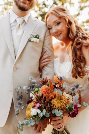 Bride with red hair leans on groom holding stunning autumn bouquet with protea and eucalyptus.