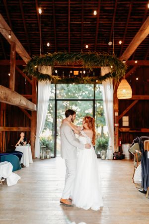 Bride and groom sharing first dance in rustic barn venue with string lights and greenery chandelier.