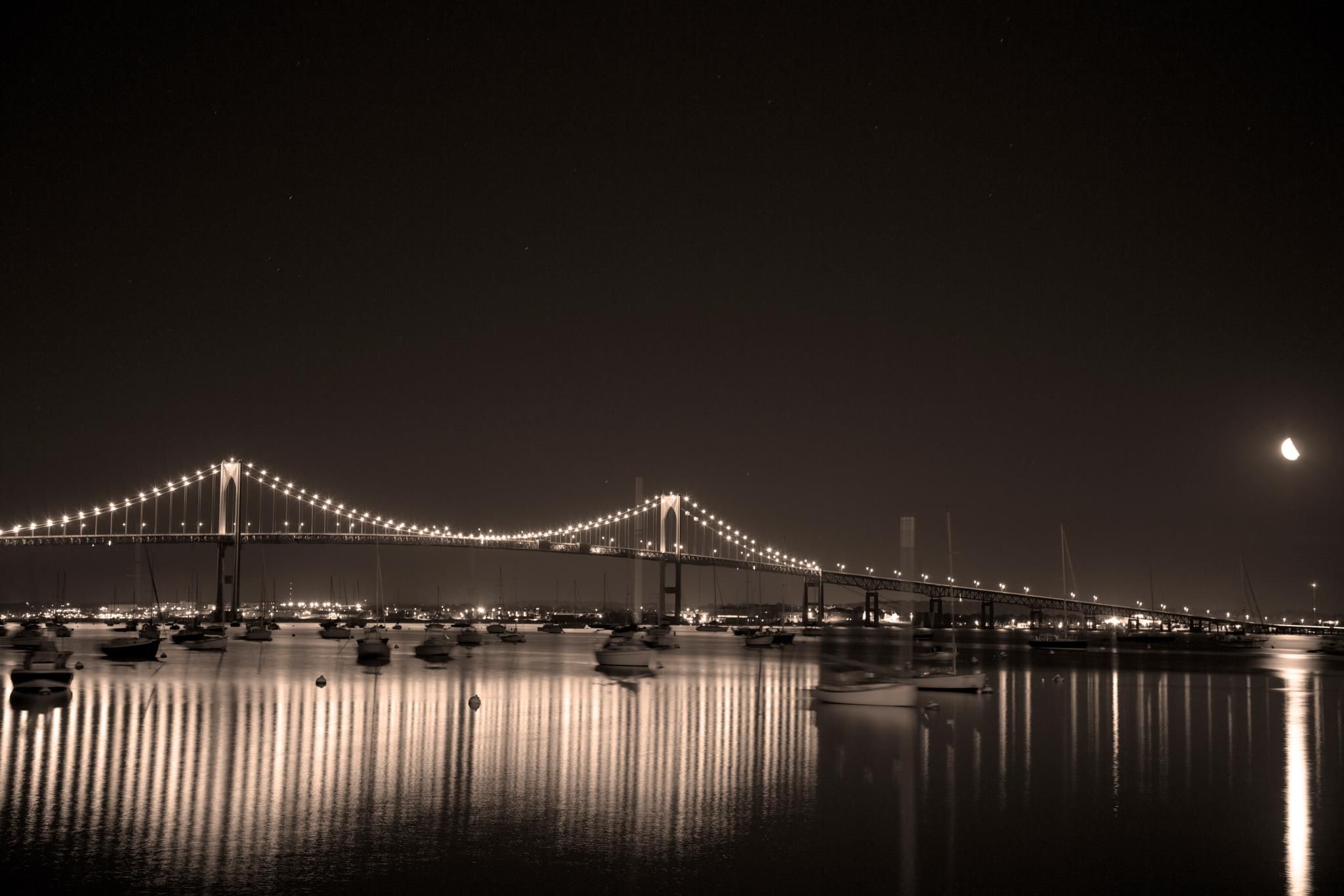 Illuminated suspension bridge reflecting in calm water at night with moon in the sky.