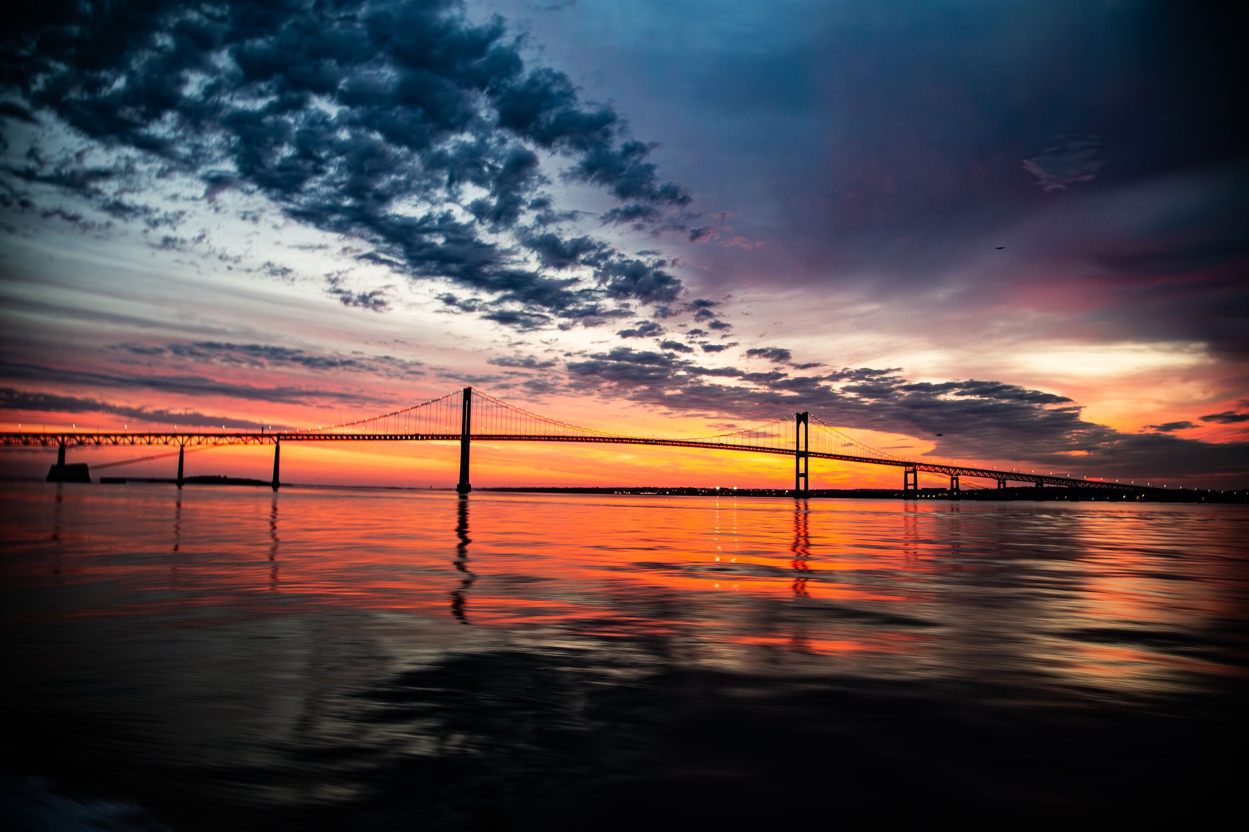 Dramatic orange sunset over a bridge silhouetted against colorful clouds reflecting on calm water.
