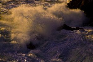 Dramatic ocean waves crashing against rocks at sunset creating golden mist.
