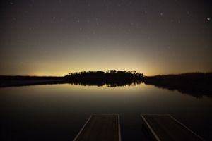 Dark lake reflection at night with wooden dock silhouetted against starry sky.
