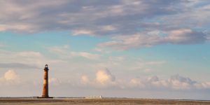 Morris Island lighthouse stands tall against pastel sky with puffy clouds on the horizon.