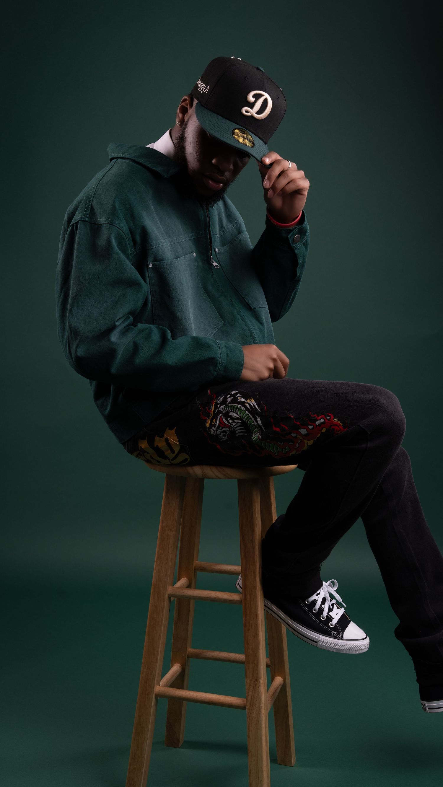 Stylish person in dark hoodie and baseball cap sitting on wooden stool against green background.