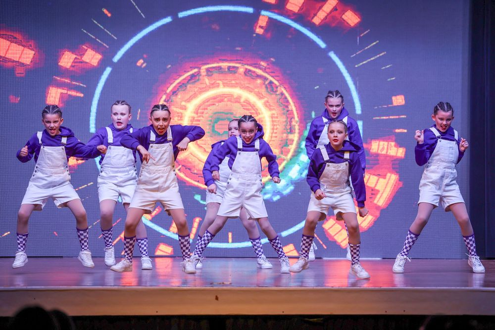 Dancers in white uniforms perform synchronized moves on stage with a glowing circular neon backdrop.