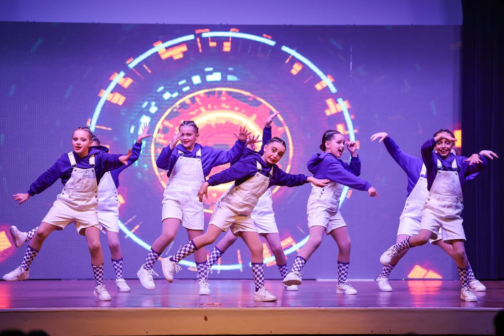 Dance group leaps in unison wearing matching white outfits against a colorful circular light display.