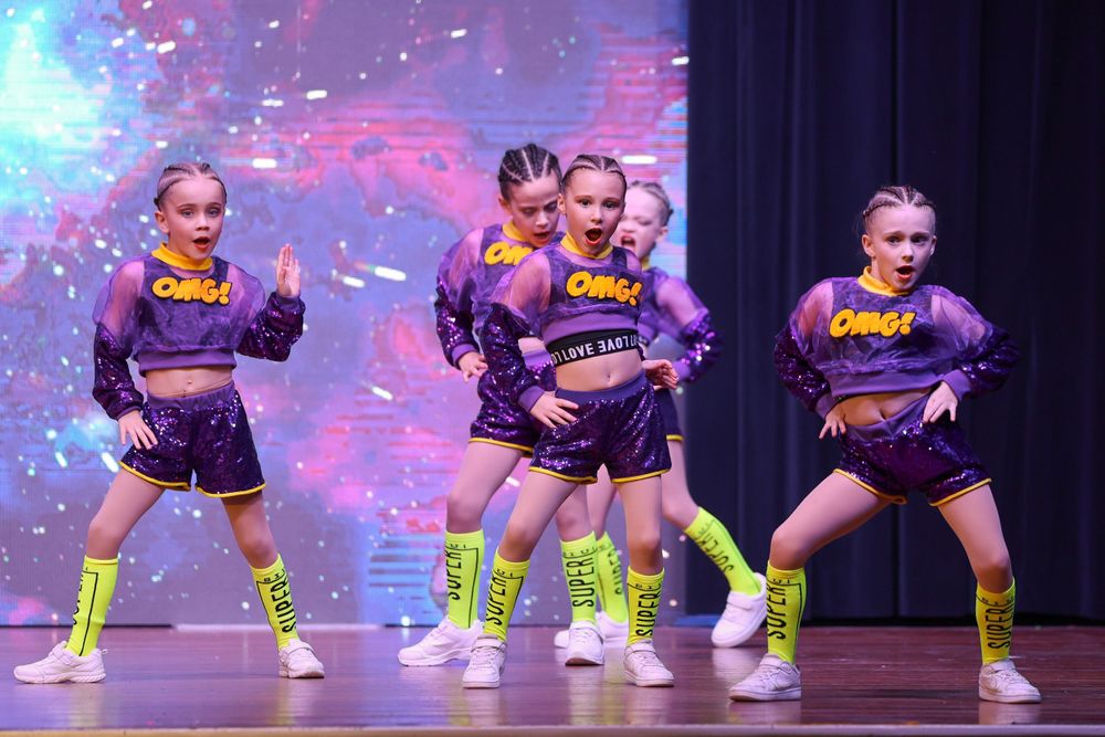 Dancers in purple outfits and neon yellow boots perform energetic choreography on stage.
