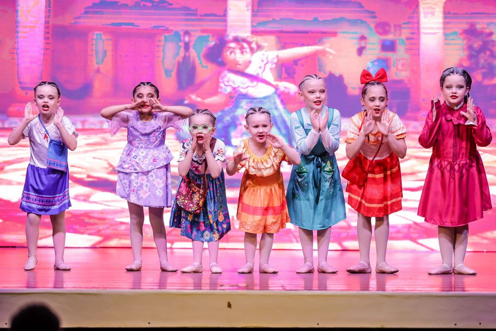 Young dancers in colorful dresses perform on stage with a pink backdrop.