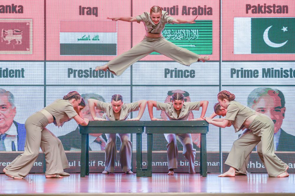 Modern dancers perform against backdrop displaying international flags and leader portraits.