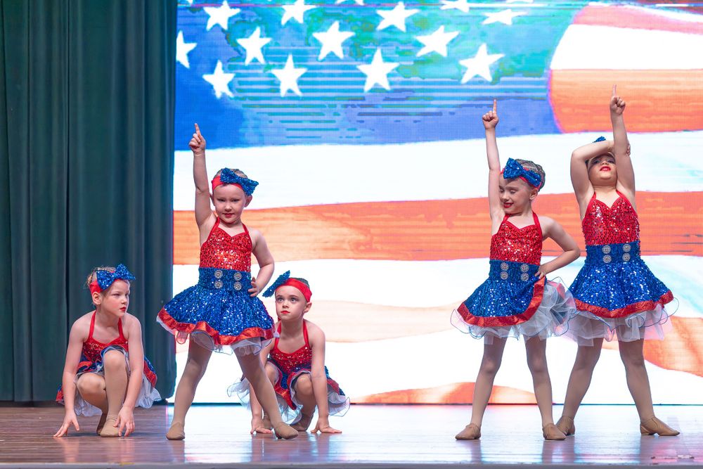 Dancers in patriotic red white and blue costumes perform in front of an American flag backdrop.