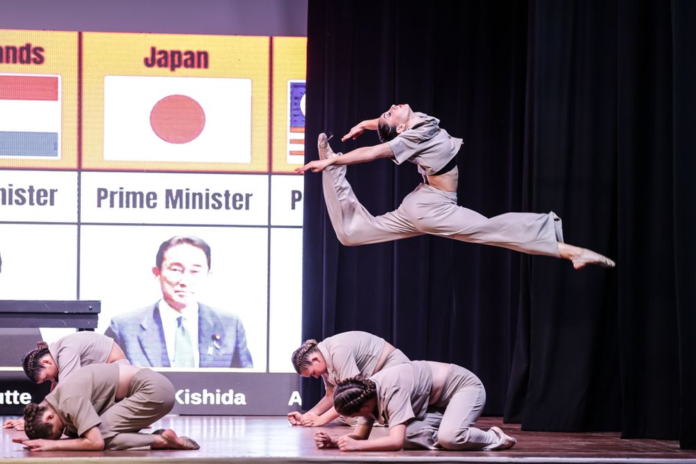 Dramatic dance performance with leaping figure and background display showing international leaders.