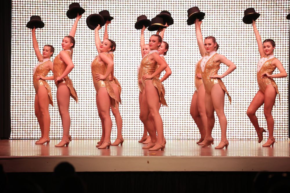Performers hold black hats while posing in matching nude-colored costumes on a theatrical stage.