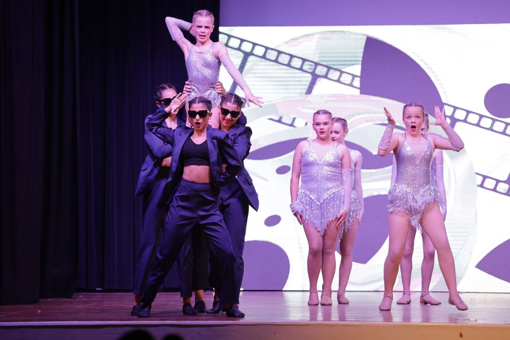 Performers in black and silver costumes dance in formation against a film-themed backdrop.