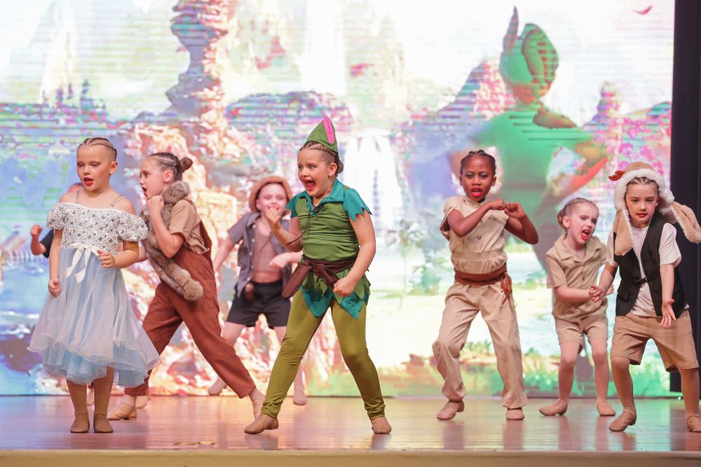Children perform a whimsical dance number in costume against a fantasy-themed digital backdrop.
