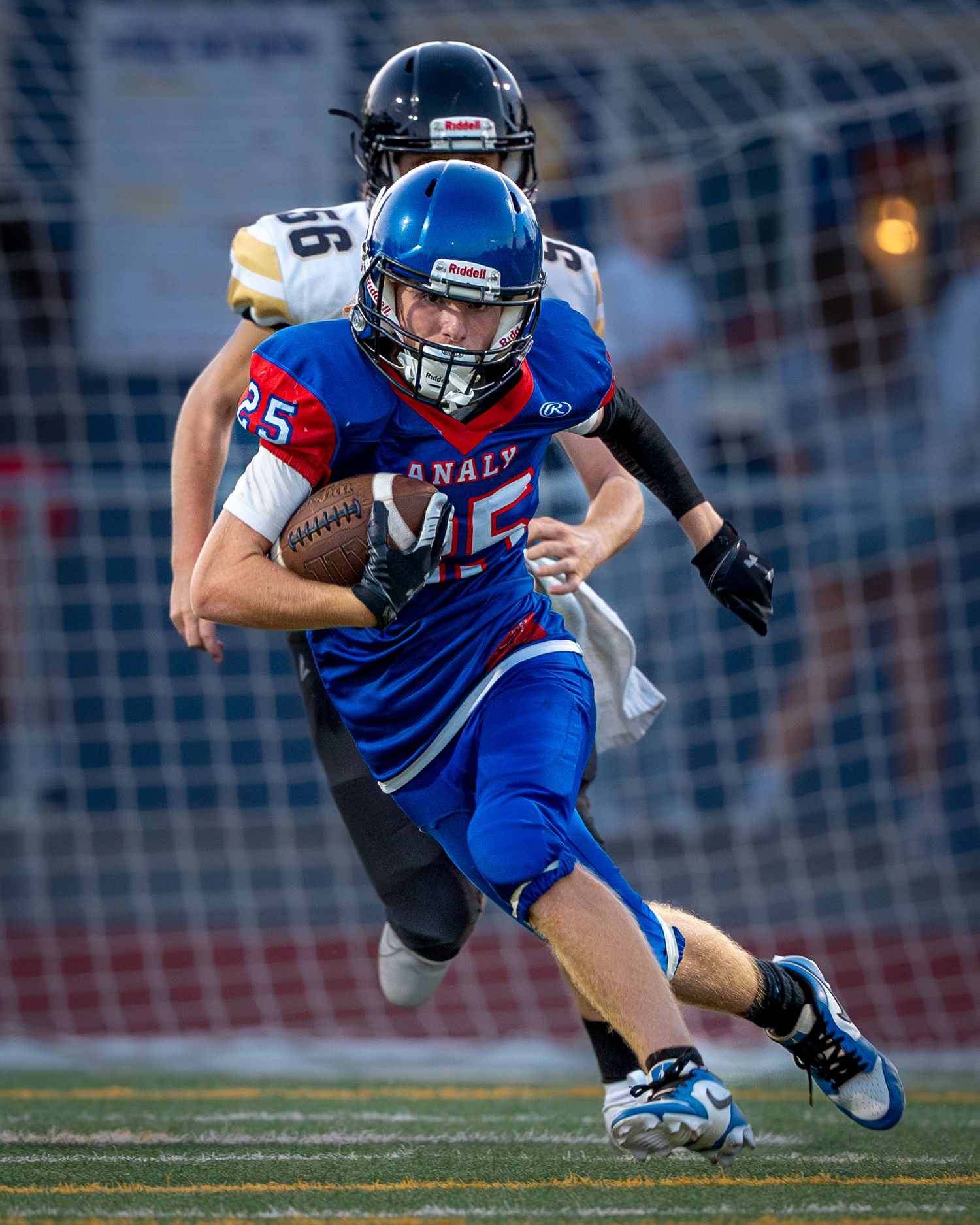 Football player in blue jersey carries ball while running past defenders during night game.