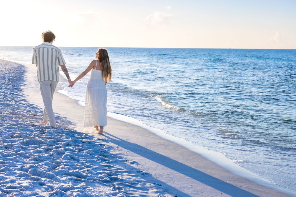 A couple holds hands while walking along a pristine beach shoreline with blue ocean waves at sunset.