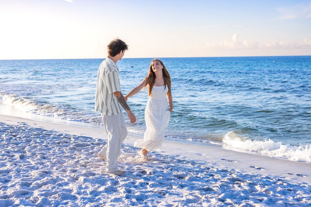 A couple in white attire walks hand in hand along a pristine beach with turquoise waters and gentle waves.