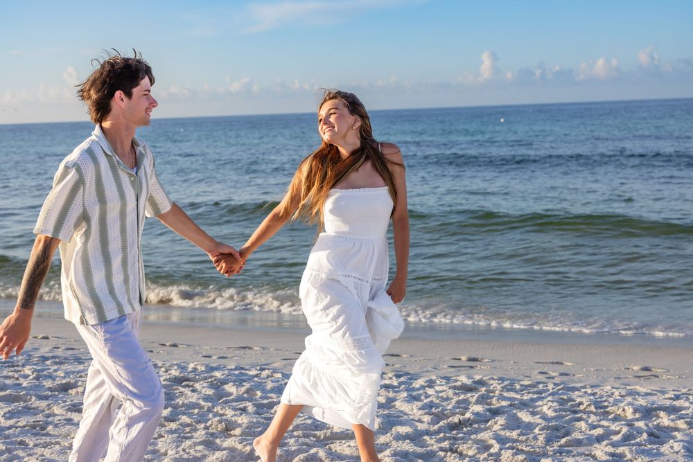 A joyful couple in white clothing runs along a sandy beach with ocean waves and blue sky in background.