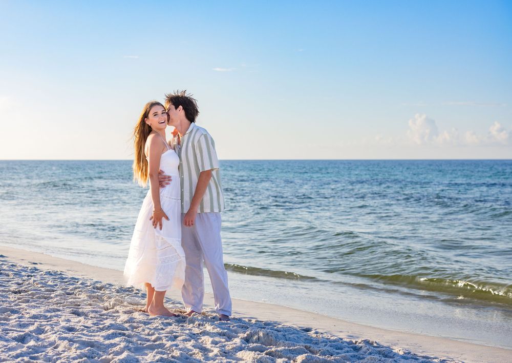 Couple embracing on white sandy beach with turquoise ocean waters and clear blue sky in background.