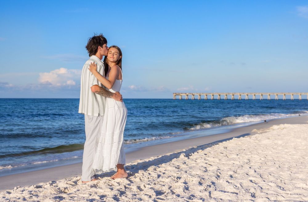 Romantic couple in white outfits walking along a pristine beach with gentle waves and blue skies.