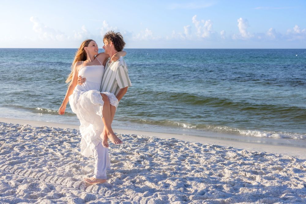 A couple dressed in white embraces while standing on a white sandy beach with turquoise ocean waters behind them.