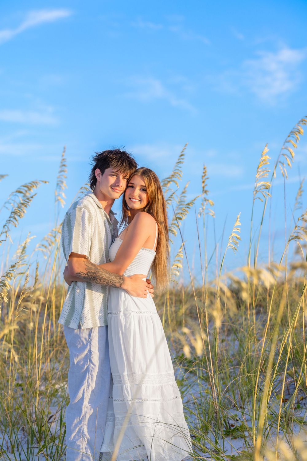 Couple in flowing white attire pose together among beach grass with golden sunlight.