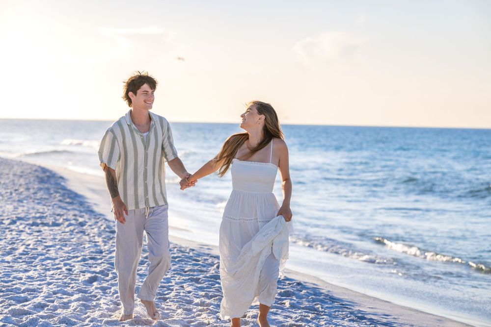 A couple walks together holding hands along a white sandy beach at sunset beside the ocean.