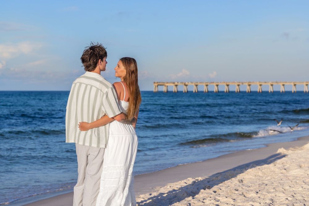 Couple strolling hand in hand on beach shoreline with pier visible in distance.
