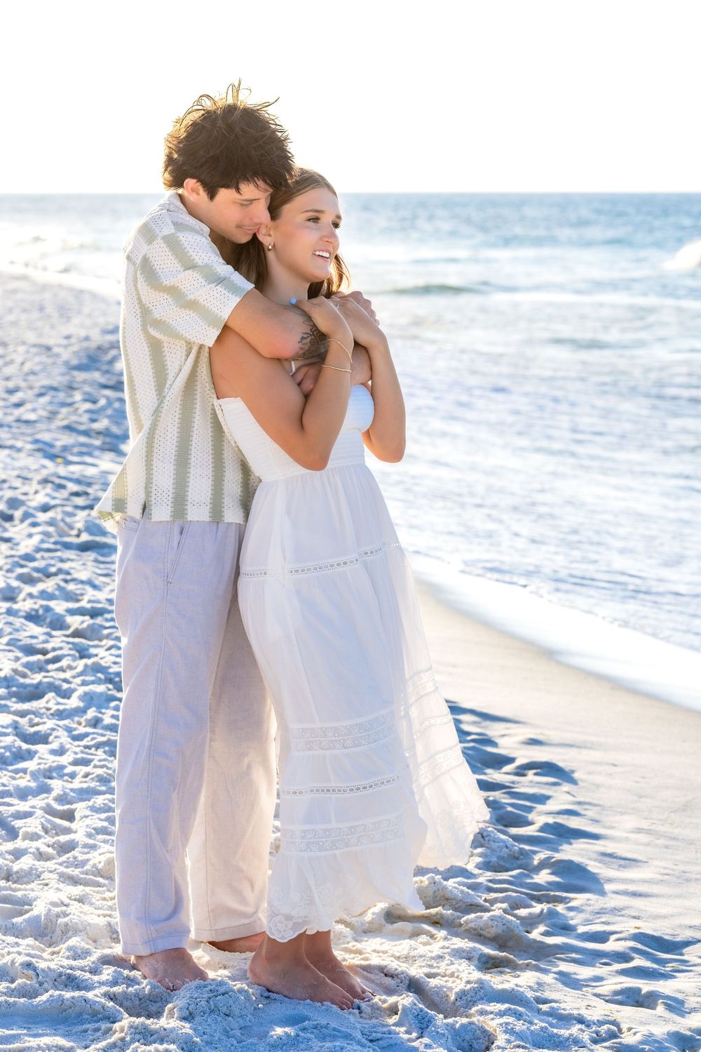 A romantic couple in white attire embrace while sitting on a sandy beach with ocean waves in the background.