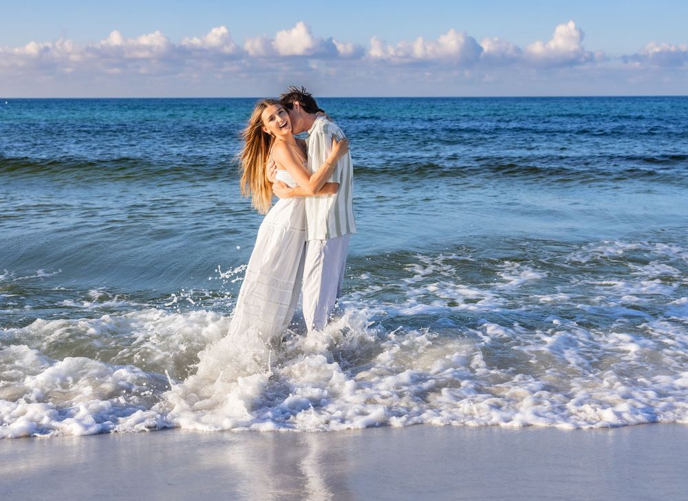 A romantic moment captured on the beach as waves crash around a couple in white attire embracing by the ocean.