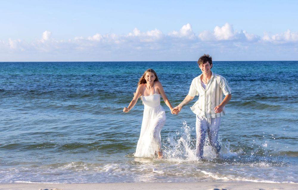 Happy couple splashes in ocean waves while wearing white beach wedding attire.
