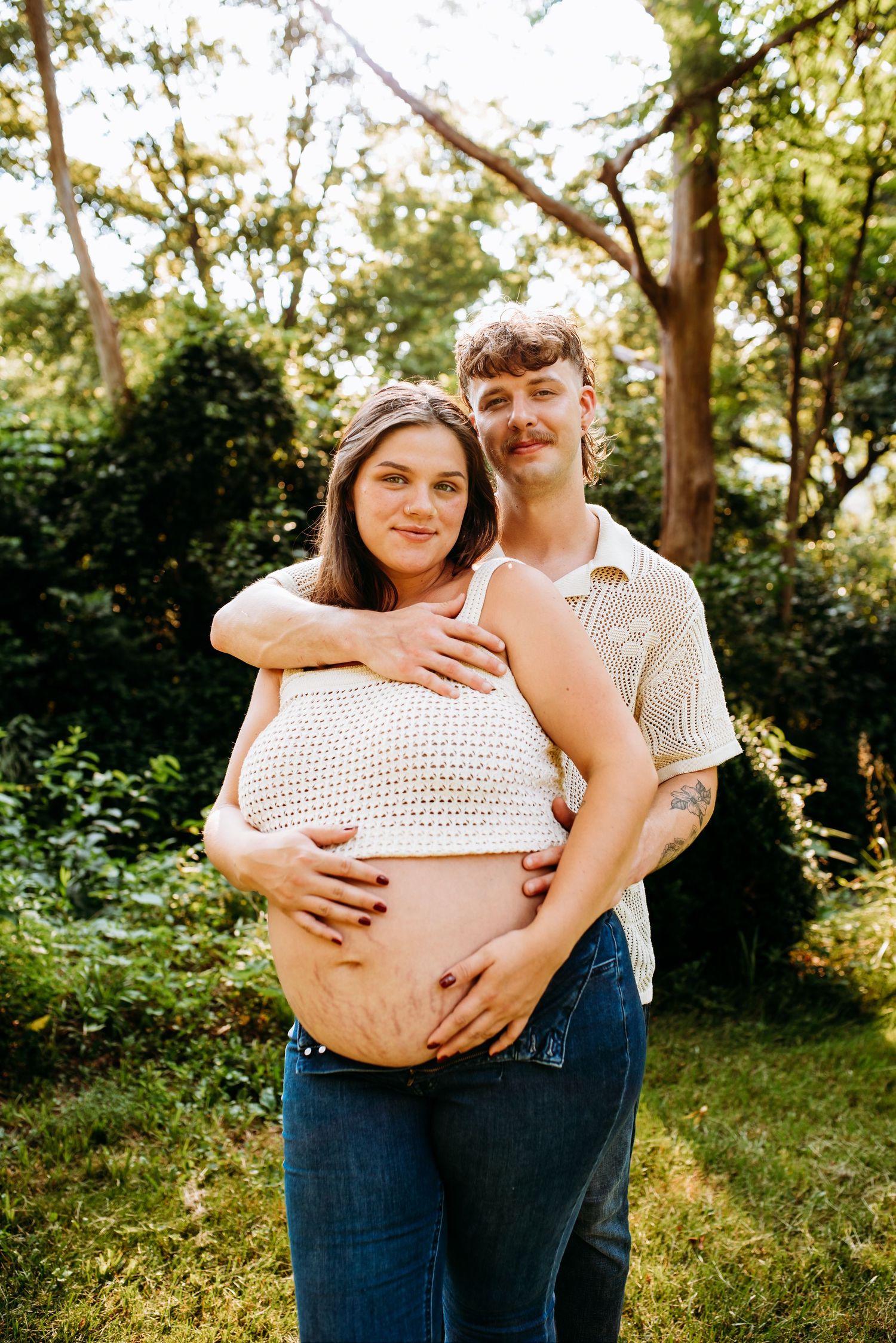 A maternity photoshoot in a sunlit garden shows an expecting couple embracing tenderly amid green foliage.