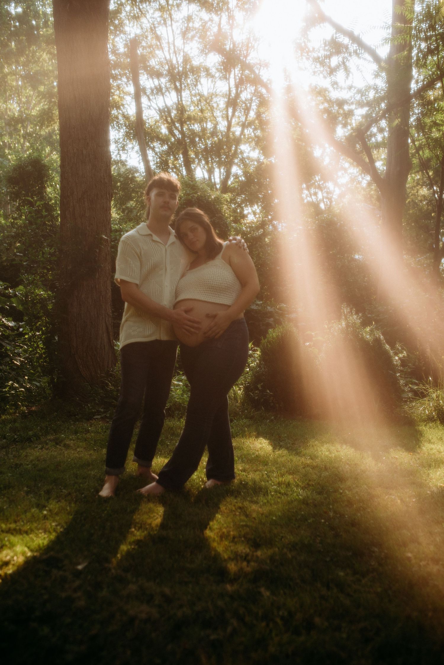 Two people share intimate moments on a grassy hill with sunbeams streaming through trees at golden hour.