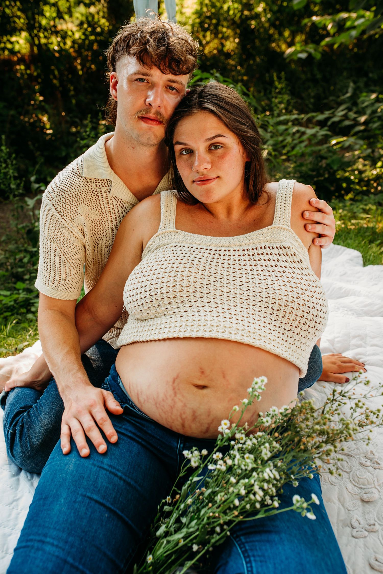 Intimate maternity portrait on a white blanket outdoors showcasing a pregnant woman in a crocheted top with her partner.